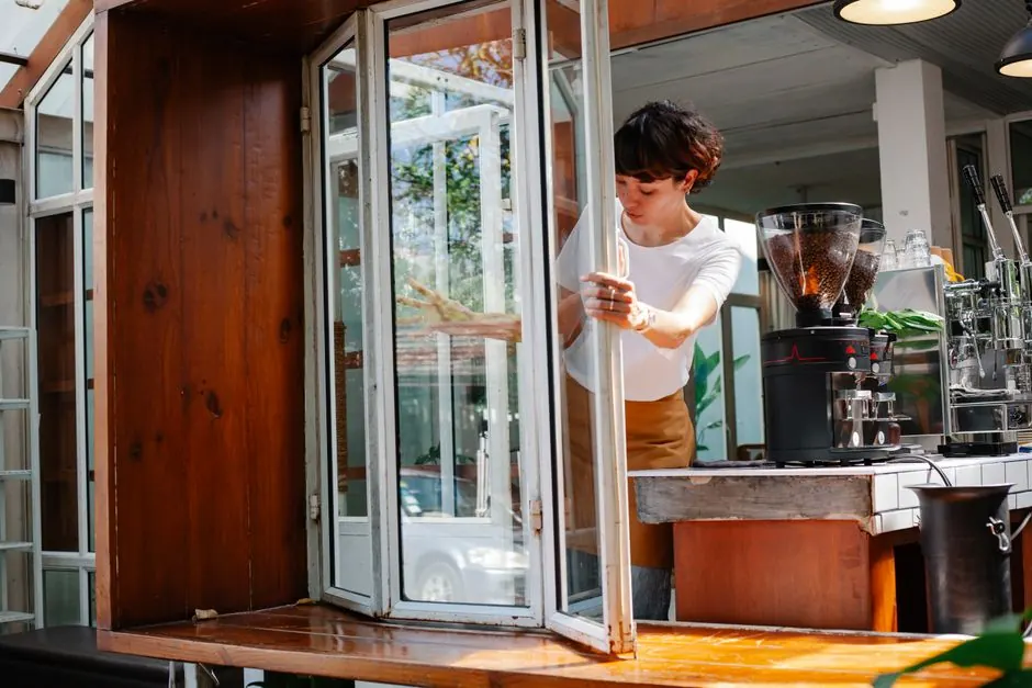 Concentrated female worker in apron opening glass window of modern coffee house with coffee machines during preparation for work day